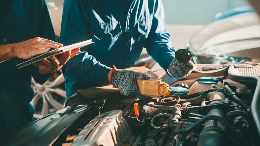 Mechanic pouring oil in car engine when his colleague using tablet computer.