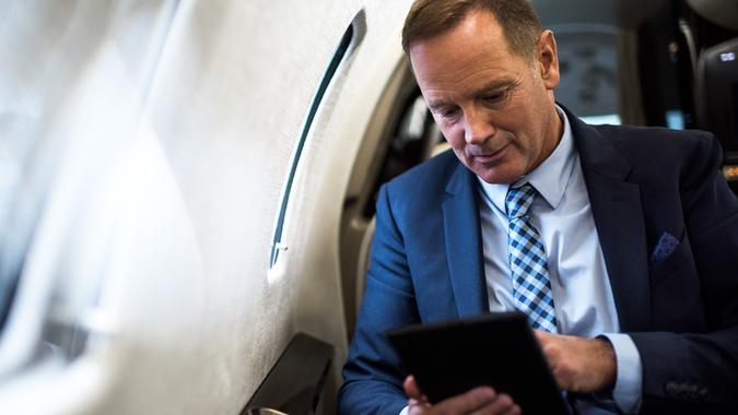 Elegant senior man sitting inside private jet airplane and holding digital tablet.