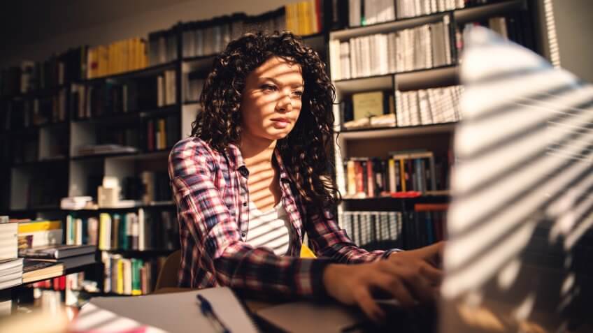 Focused young cute student girl sitting in library and looking at computer.