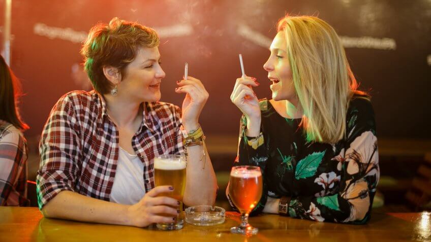 Two mid adult women having beer at the bar counter, talking and smoking cigarettes.