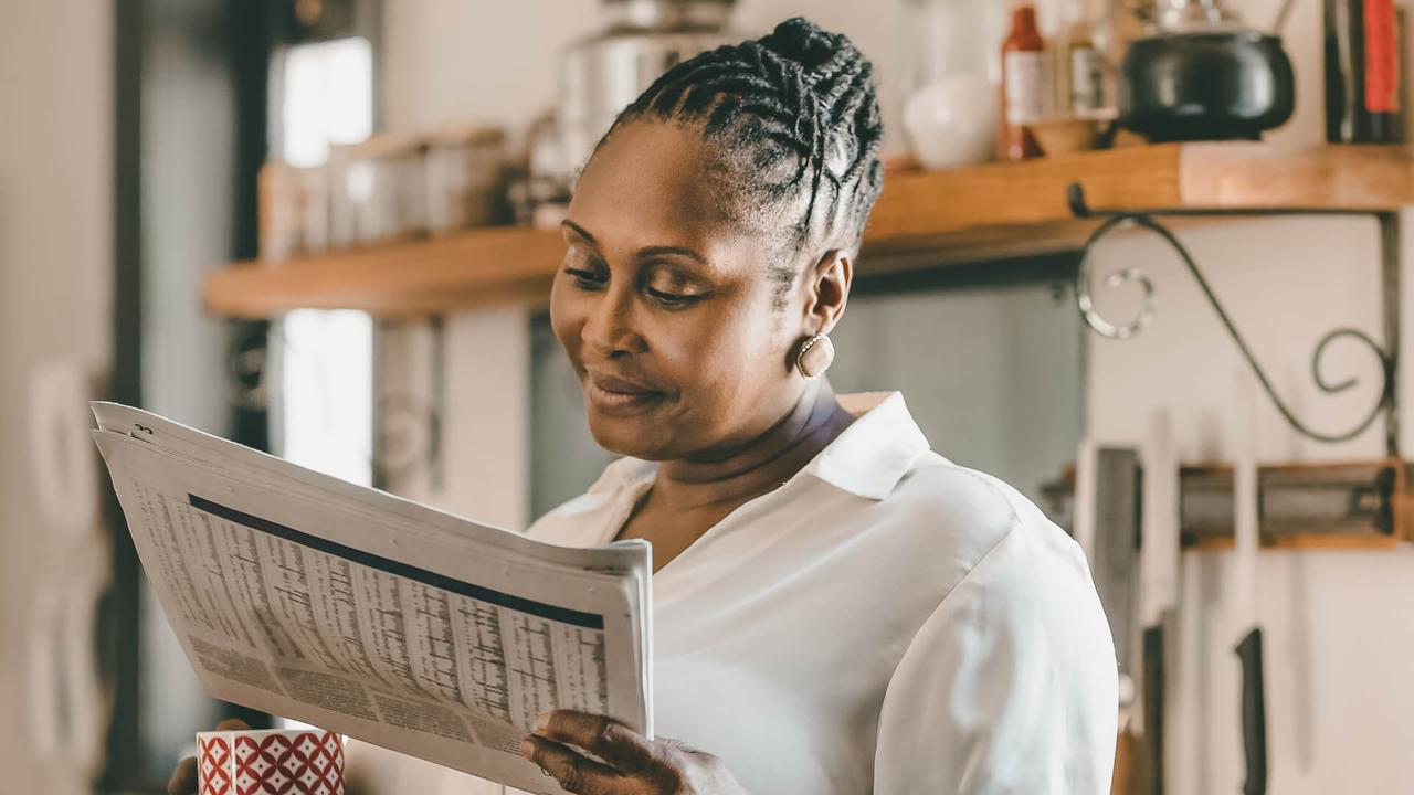 Smiling African American woman drinking a cup of coffee and reading the newspaper while standing in her kitchen in the morning.