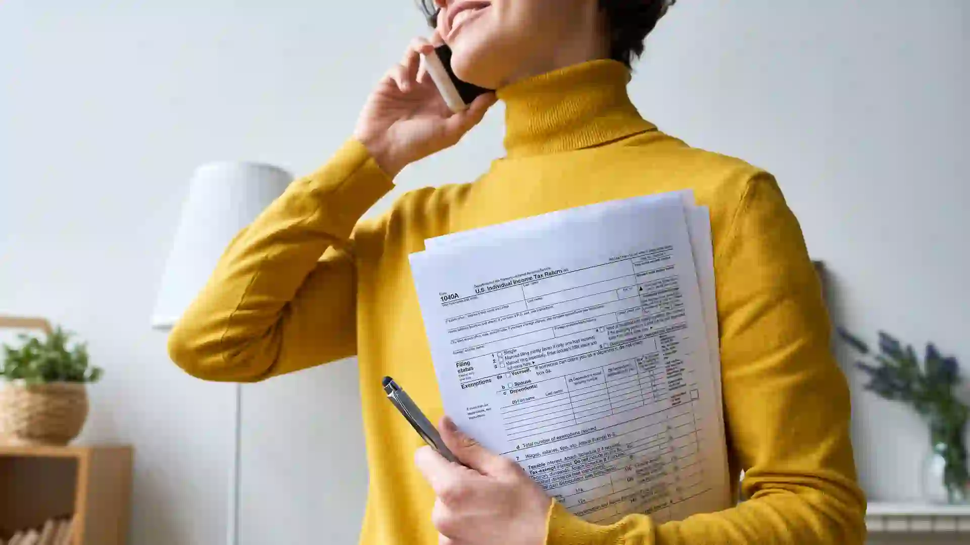 Close-up of positive young woman in glasses standing in living room and holding individual income tax return form while talking by mobile phone.