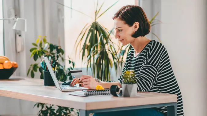 Woman using laptop and shopping online while holding credit card at the desk by the window.