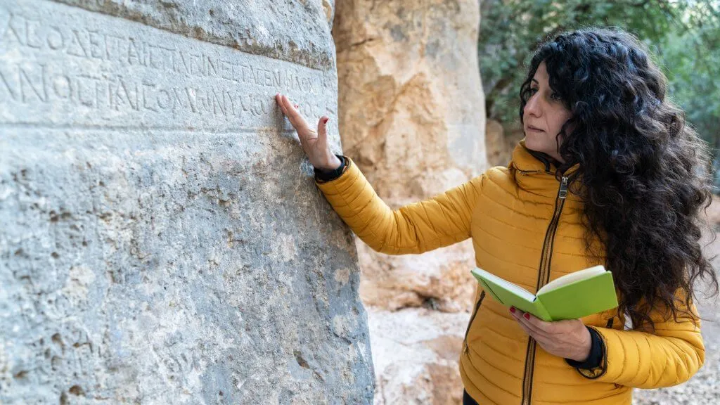 Female archaeologist standing by a stone antique stone structure and writing notes to notebook while examining ancient Latin script written on rock.