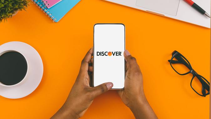 Person's hands holding smartphone with Discover app open against orange background.