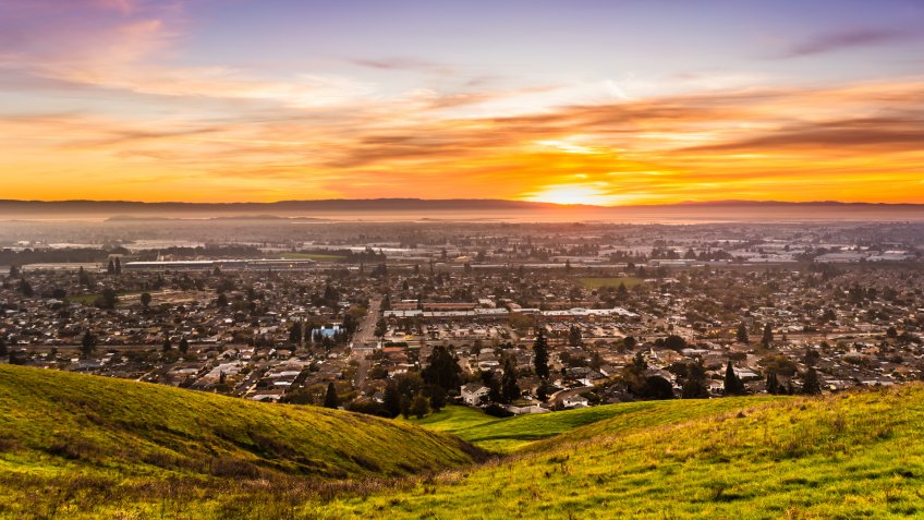 Sunset view of residential and industrial areas in East San Francisco Bay Area; green hills visible in the foreground; Hayward, California.