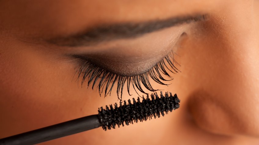 closeup of a young dark-skinned woman applying mascara on a white background.