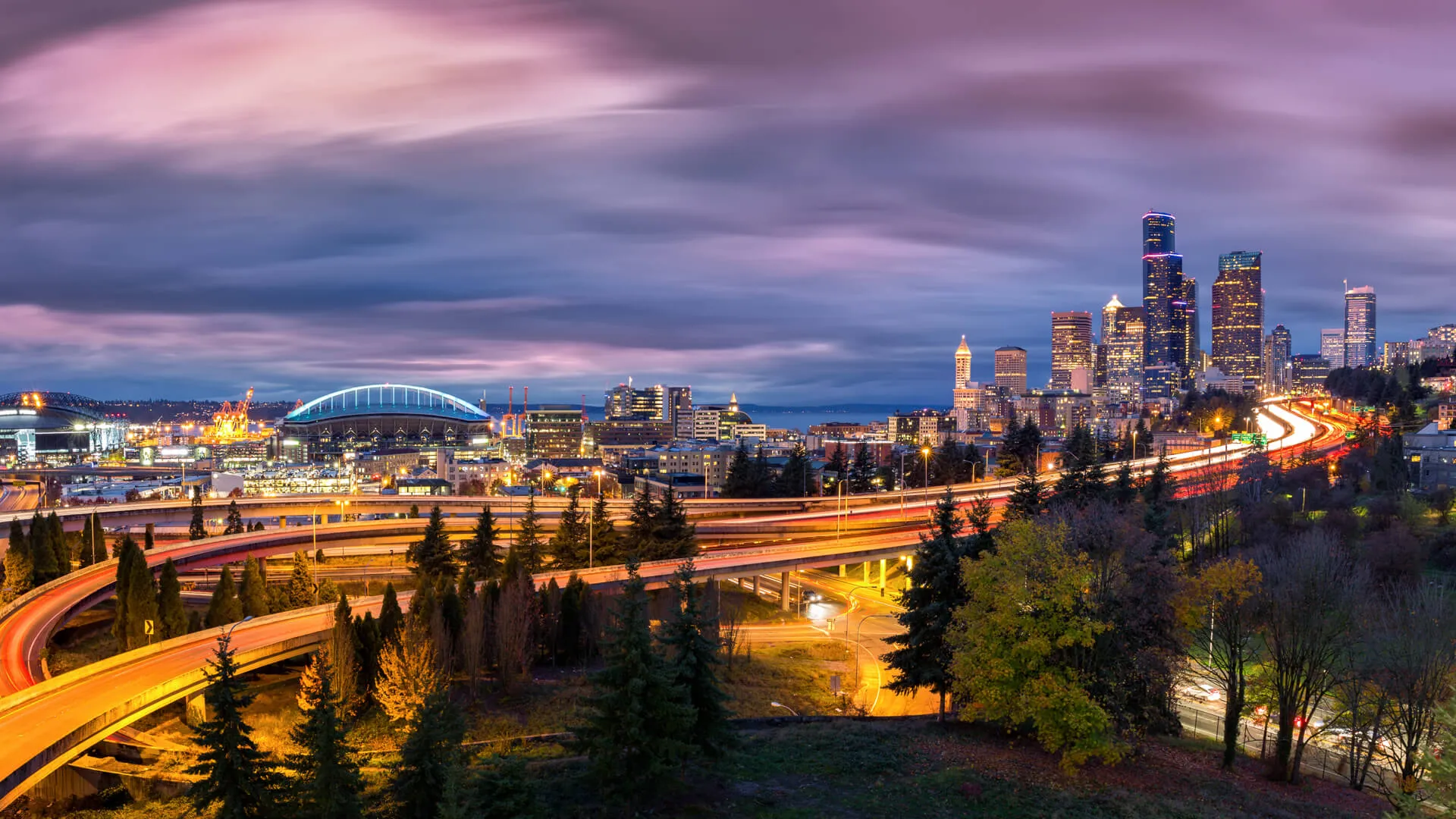 Seattle cityscape at dusk with skyscrapers, winding highways parks and sports arenas under a dramatic sky.