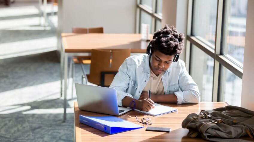 African American teenage boy writes something in a notebook while studying in the campus library.