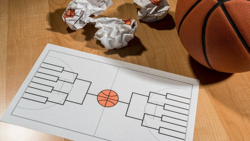 Attempting to fill out a college basketball tournament bracket on a legal size piece of paper with several crumpled balls of paper with failed attempts, sitting on a basketball court with a basketball sitting next to it.