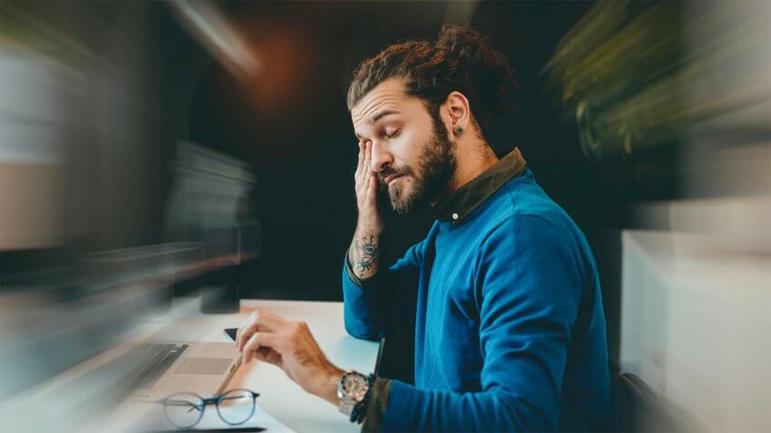 Tired bearded hipster sitting in his office and rubbing his eyes.