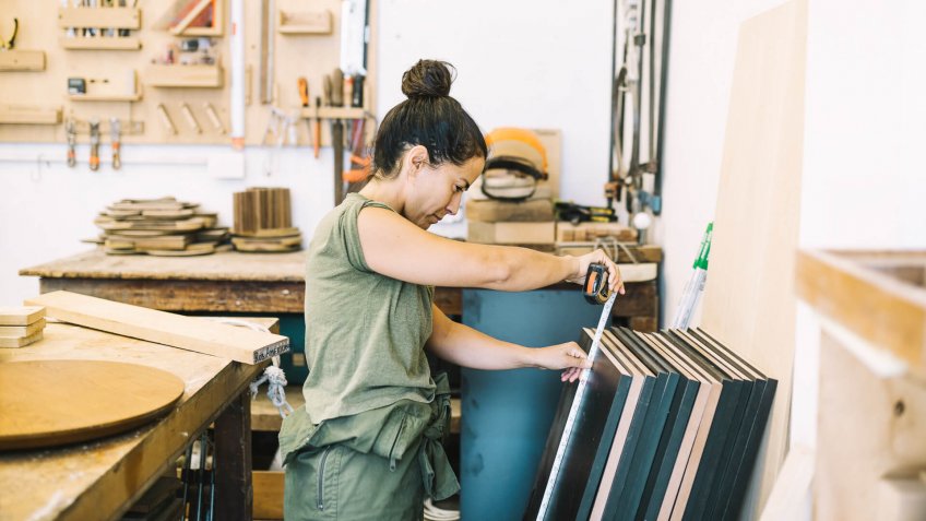 Candid portraits of young woman working in a wood workshop - taking measurements on a piece of wood.