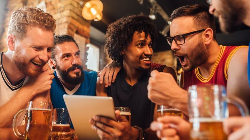 Multi - ethnic group of basketball fans watching a basketball game on the tablet and drinking beer at the pub.