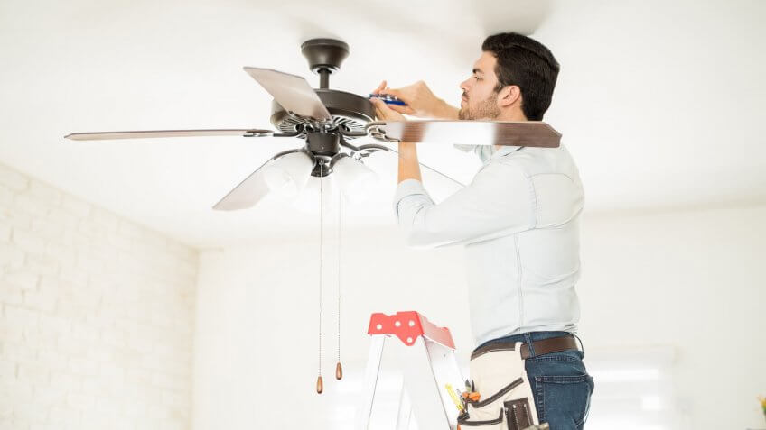 Attractive young handyman stepping on a ladder and fixing a ceiling fan.