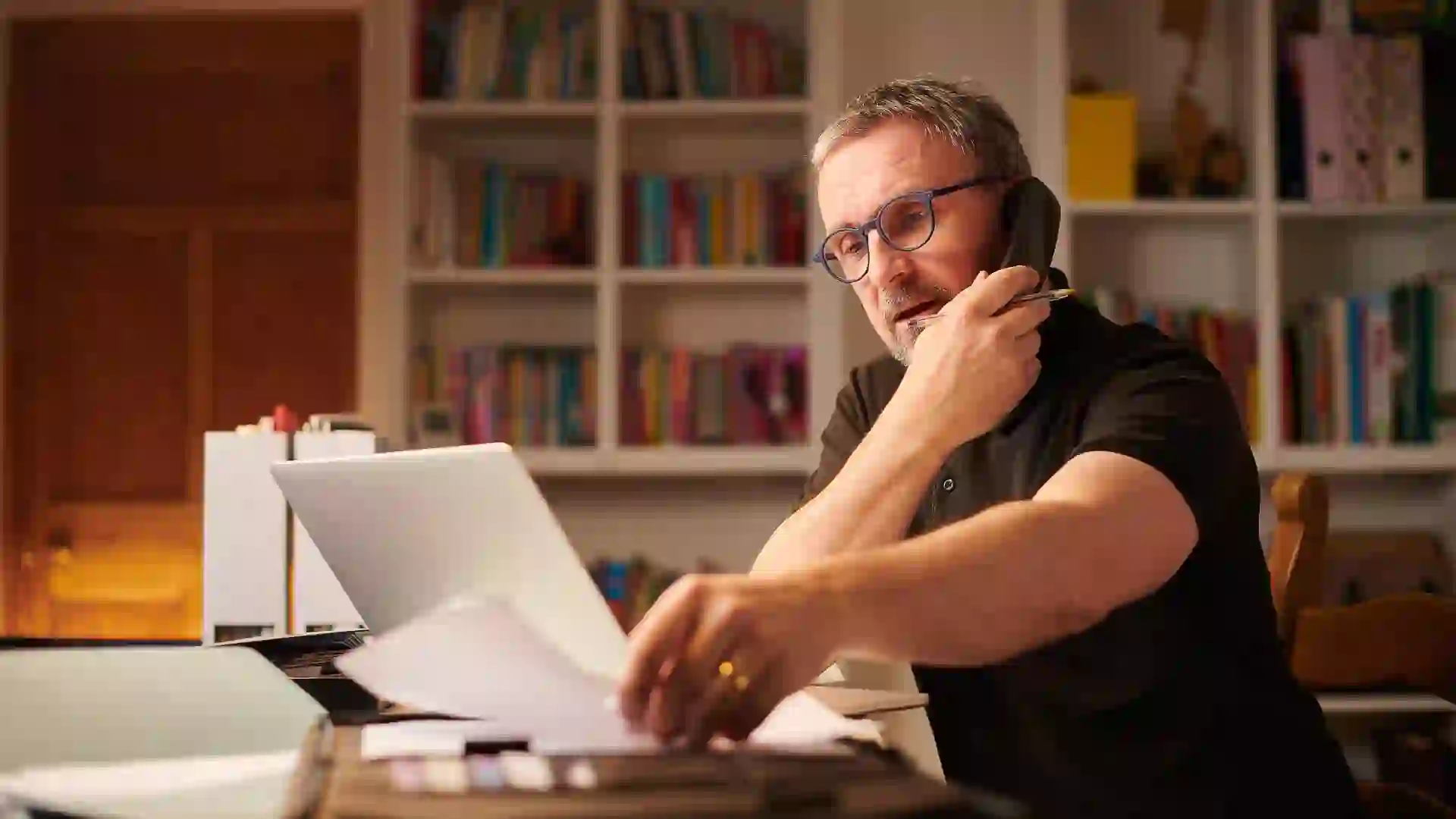 A mature man is working from home or sorting the home finances, with paperwork and a laptop.