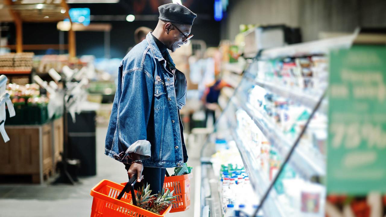 Stylish casual african american man at jeans jacket and black beret holding two baskets, standing near fridge and shopping at supermarket.