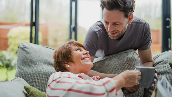 Man is leaning over the sofa to give his mother a cup of tea.