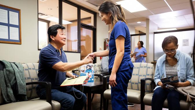 A mid adult female nurse shakes hands with the mature adult male patient in the busy waiting room.