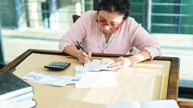 Senior female accountant calculating finances Taiwanese senior woman sitting at table, using calculator and writing results on paper.