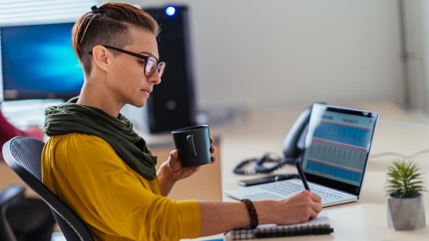 Businesswoman wearing yellow shirt writing notes in the office and drinking coffee.