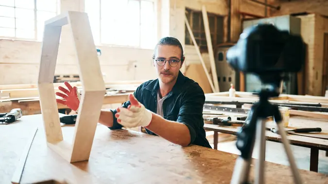 Shot of young carpenter recording a tutorial of him making a wooden masterpiece on a video camera inside his workshop.