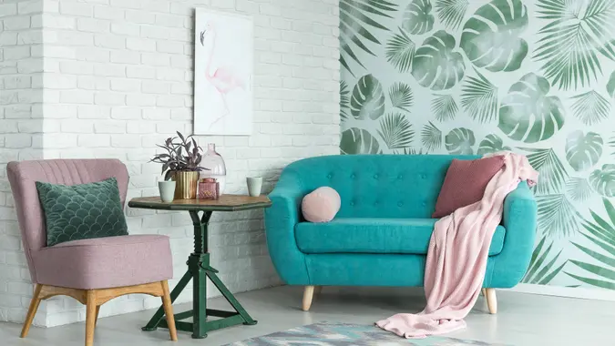 Green table with plant between pink chair and blue sofa in floral living room with wallpaper and poster.
