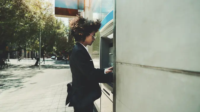 Side view of curly man in foraml suit using street ATM to withdraw cash from his account; a businessman is using an outdoor cash dispenser to increase his account.