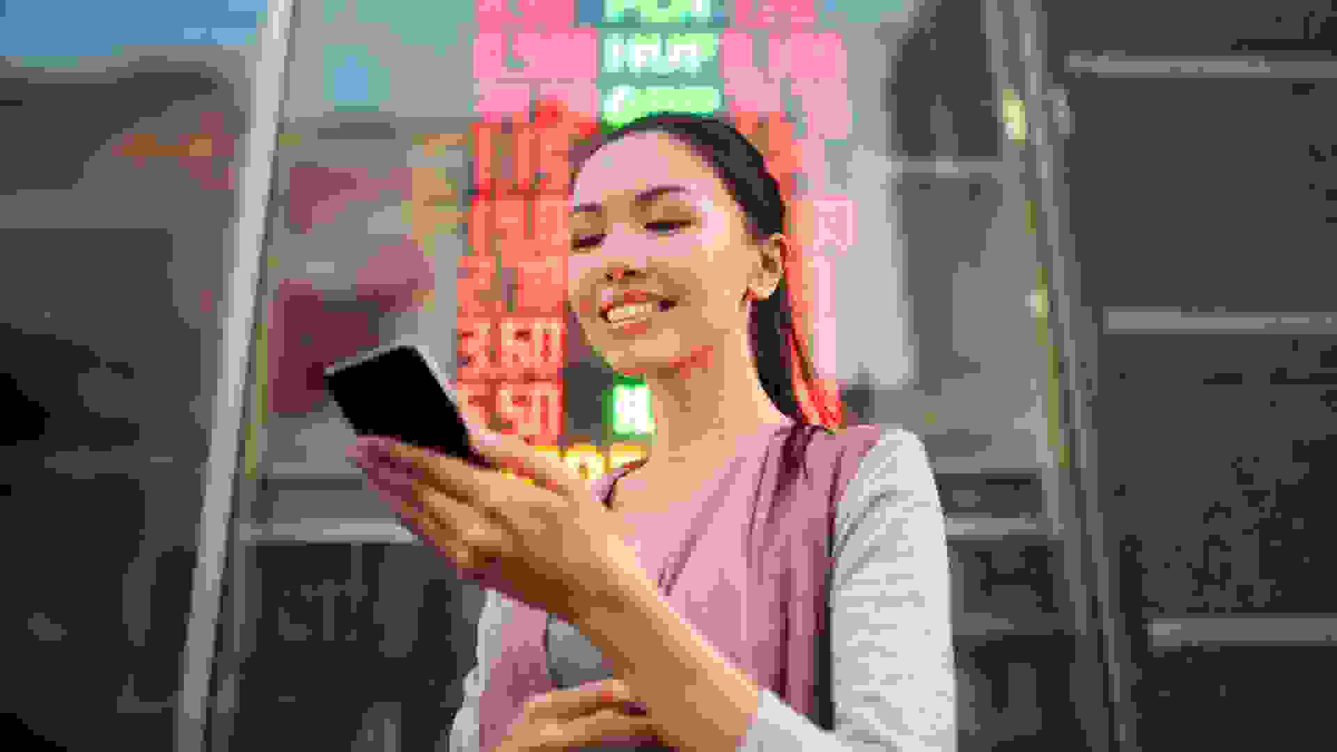 A young beautiful Asian woman using an application in her smart phone to check currency exchange rates in front of an illuminated information board.