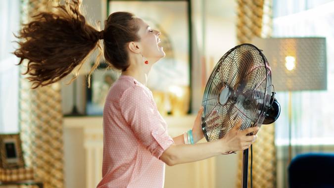 smiling housewife enjoying fresh air in front of working fan smiling modern housewife in the modern living room in sunny hot summer day enjoying fresh air in the front of working fan.