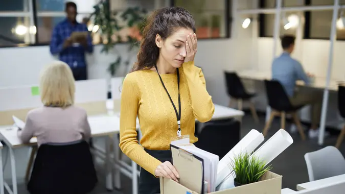 Frustrated young woman in yellow sweater standing at table and touching face with hand while packing stuff in office after dismissal.