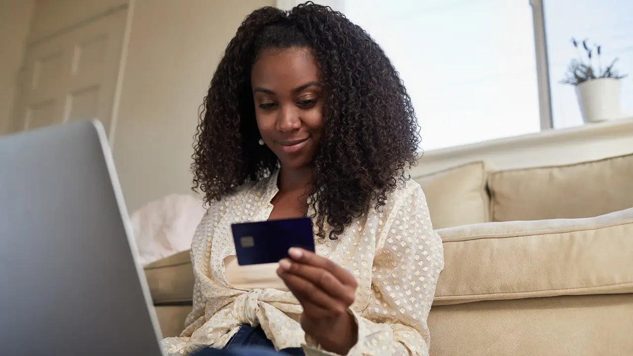 Smiling young African American woman doing some online shopping with a credit card and laptop while sitting on her living room floor.