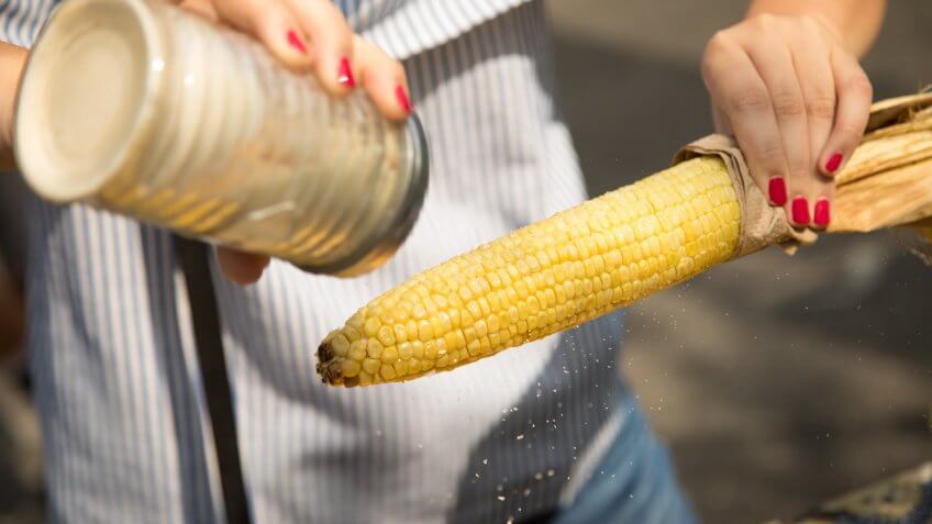 Close up on a woman holding roasted yellow Mexican corn, shaking seasoning with a canister filled with garlic chili salt on the end of the cob.