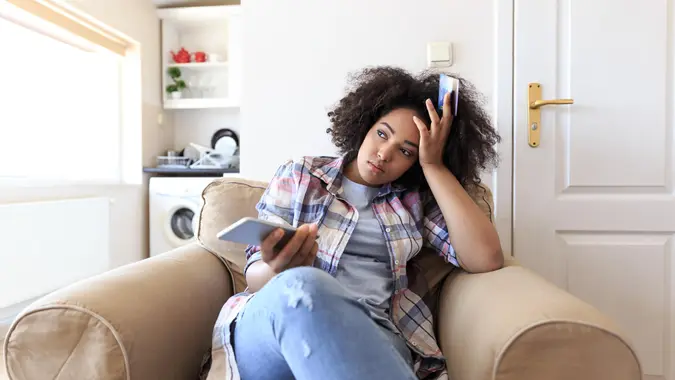 Young woman sitting at home, holding smart phone, having problems with credit card.