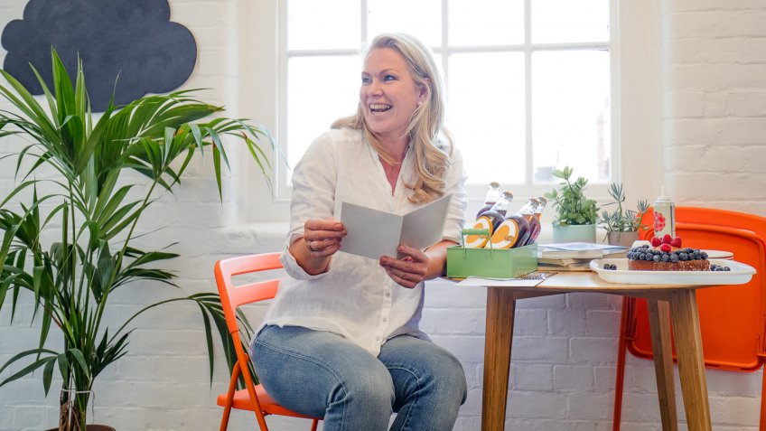 A mid adult woman reads a birthday card she has received from her colleagues, a birthday cake can be seen on the table beside her.