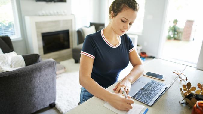 Shot of a young woman using a laptop and writing in a notepad while working at home.