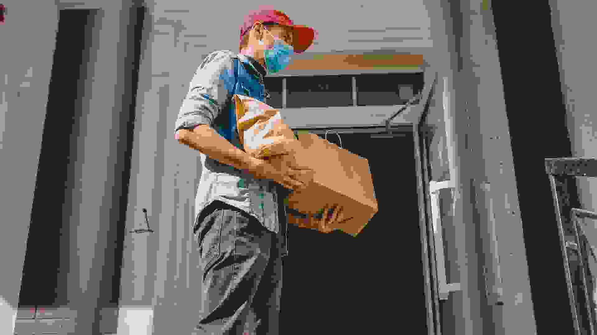 Delivery man holding paper bag with food on white entrance of house background , food delivery man in protective mask.