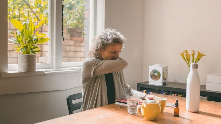 Middle aged woman sitting at table coughing into elbow - coronavirus self isolation concept (selective focus).