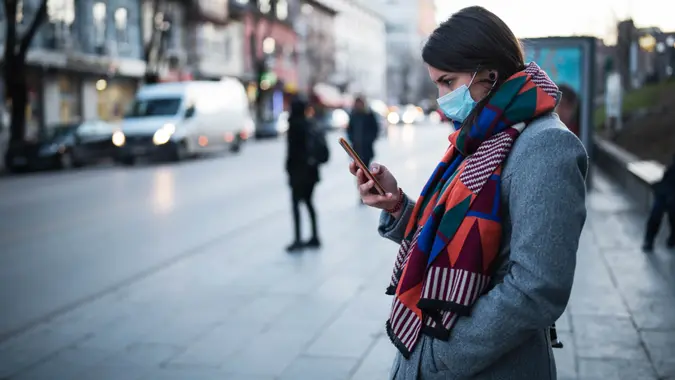 Side view of young woman with face protective mask on the street, she waiting for city transport and looking down, she using smartphone.