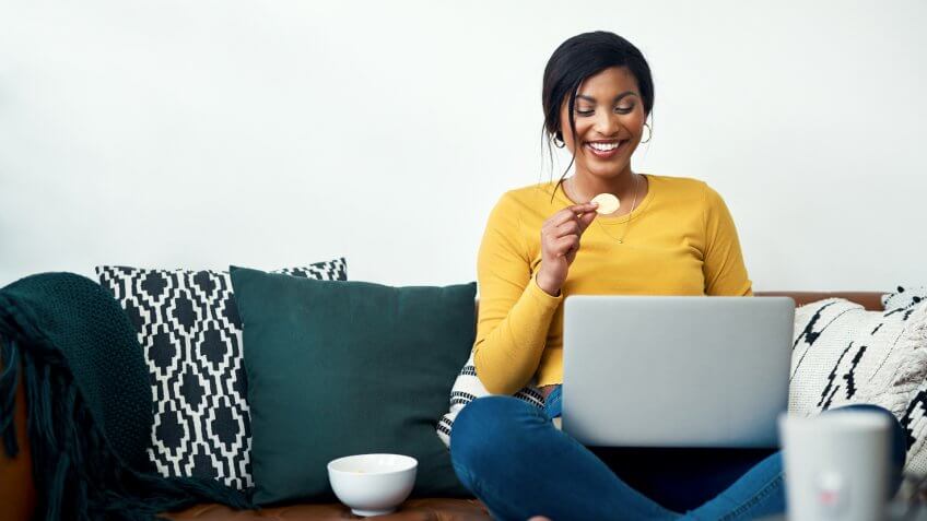 Cropped shot of an attractive young woman sitting on her sofa and eating potato crisps while using her laptop.