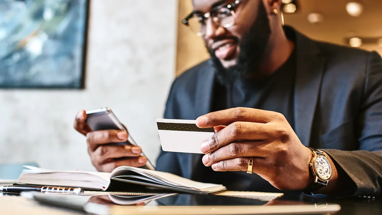 Portrait of African-American male holding cell phone in one hand and credit card in other, making transaction, using mobile banking app during lunch at cafe.