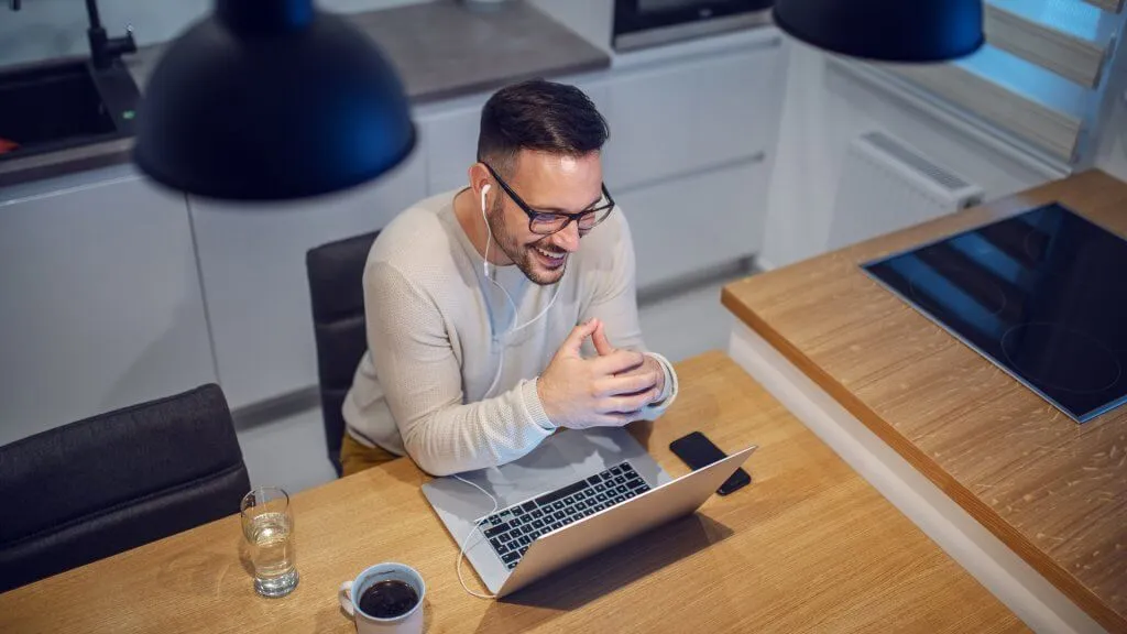 Top view of attractive smiling caucasian man sitting at dining table in kitchen and having video call with his girlfriend.
