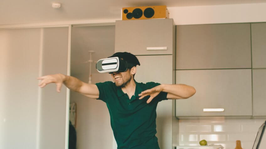 Funny young man in virtual reality 360 headset dancing in the kitchen in the morning while listening to music and have fun at home.