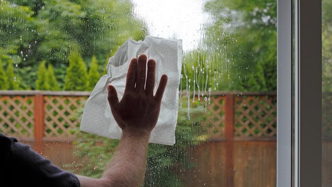Having sprayed window cleaning fluid on the glass, a hand of a man is seen washing a sliding glass door from inside a home with a view of the green foliage backyard.
