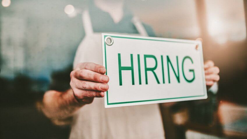 Cropped shot of an unrecognizable male coffee shop owner hanging a "HIRING" sign in his window.