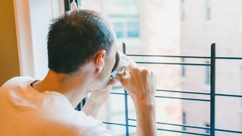 Closeup of young man drinking coffee, tea from cup looking outside of small apartment window in New York City NYC urban Bronx, Brooklyn brick housing, guard rail, security bars.