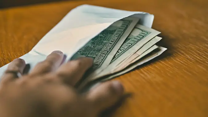 Male hand opening a white envelope full of American Dollars (USD, US Dollars) on the wooden table as a symbol of cash transfer, money laundering or bribery.