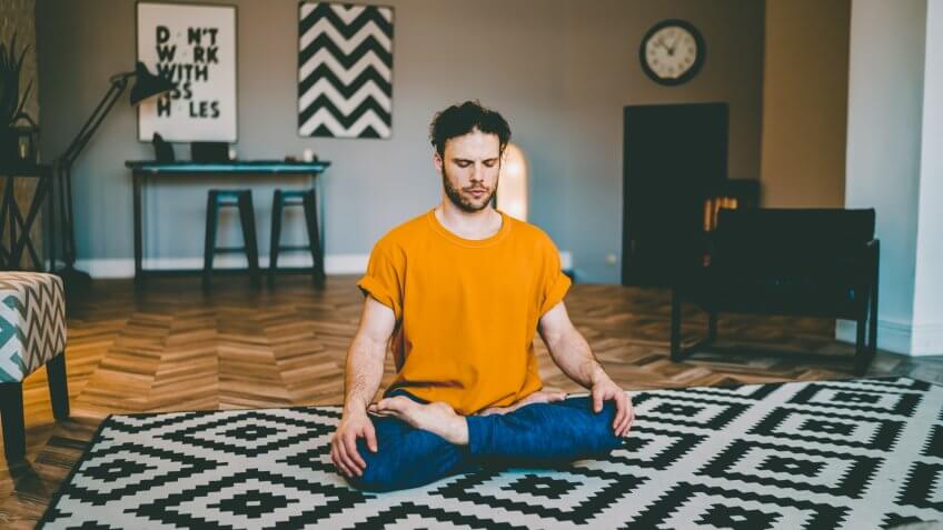 Young man with bristles sitting on yoga mat with eyes closed in center of modern living room and relaxing in easy seat pose.