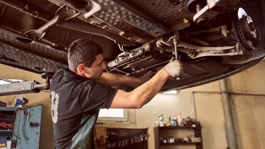 Experienced specialist car mechanic standing under lifted car during repair and maintenance process in garage.