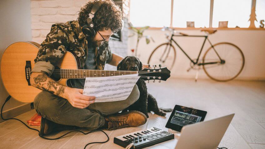 Young hipster man with tattoo sitting on floor and playing acoustic guitar, on sunny day.
