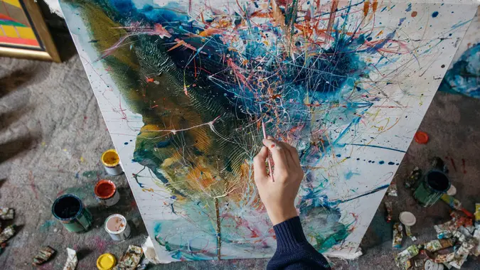 Artist- painter working on painting in studio with art supplies around him.